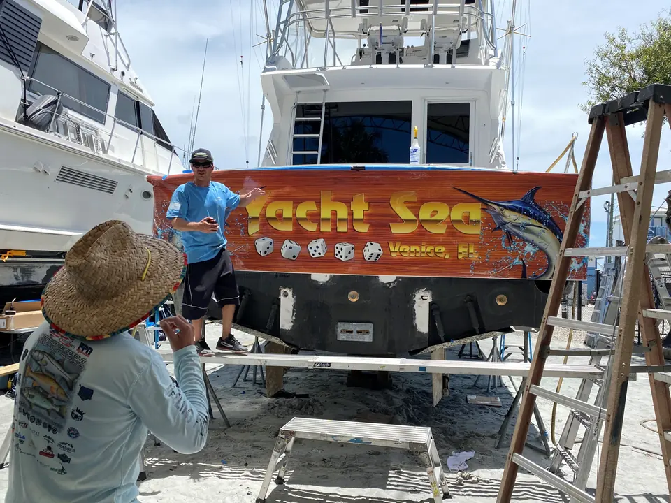 Two installers fitting the Yacht Sea transom wrap from ladders in the boatyard